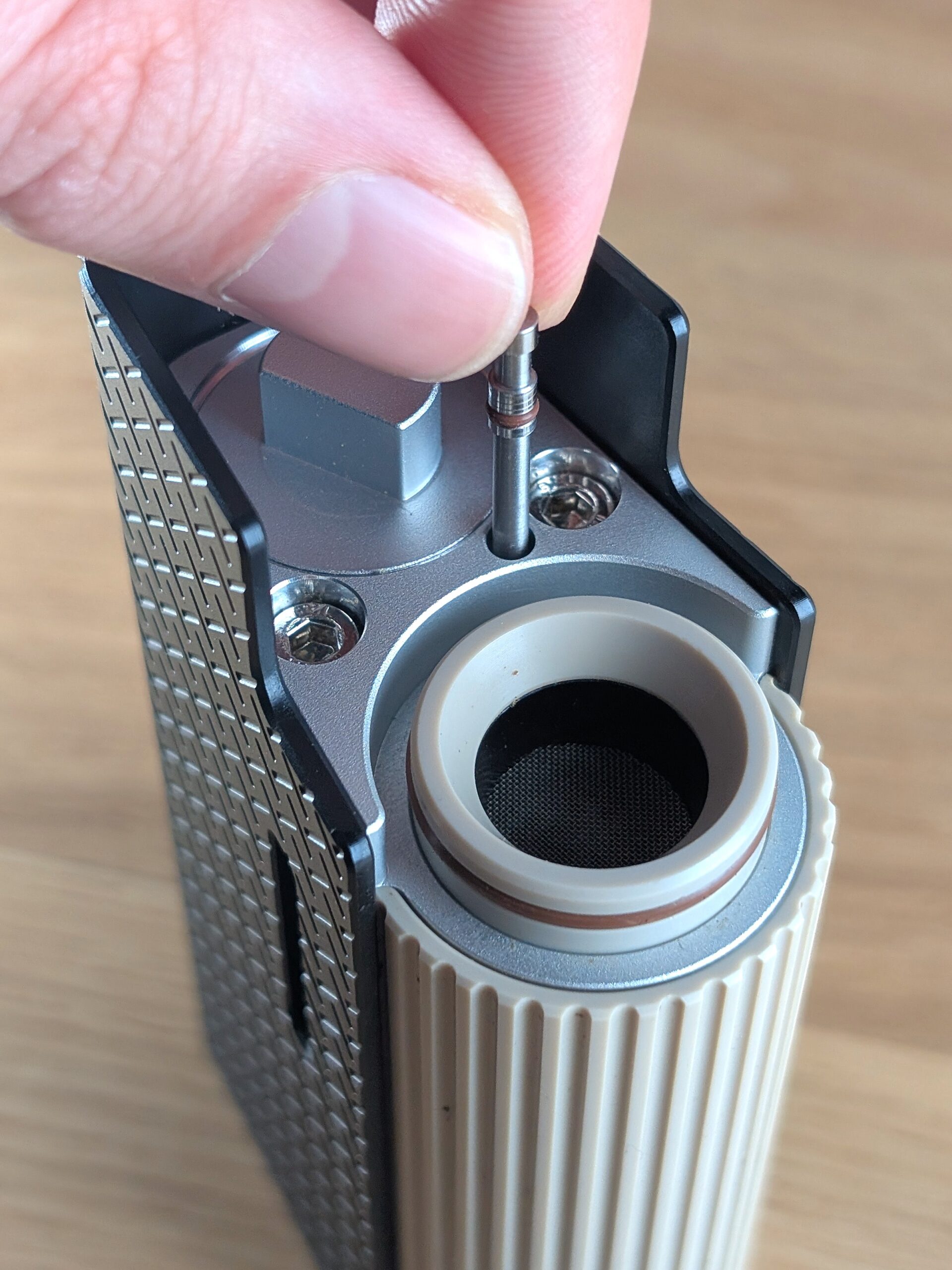 Der Frolic Thermal Extractor steht ohne Kühleinheit auf einem Holztisch. An der Obersiete des Gerätes sind Kammer, zwei Schrauben und die Batterieabdeckung zu sehen. Eine Hand zieht mit zwei Fingern das Reinigungswerkzeug aus Edelstahl aus dem Gerät.