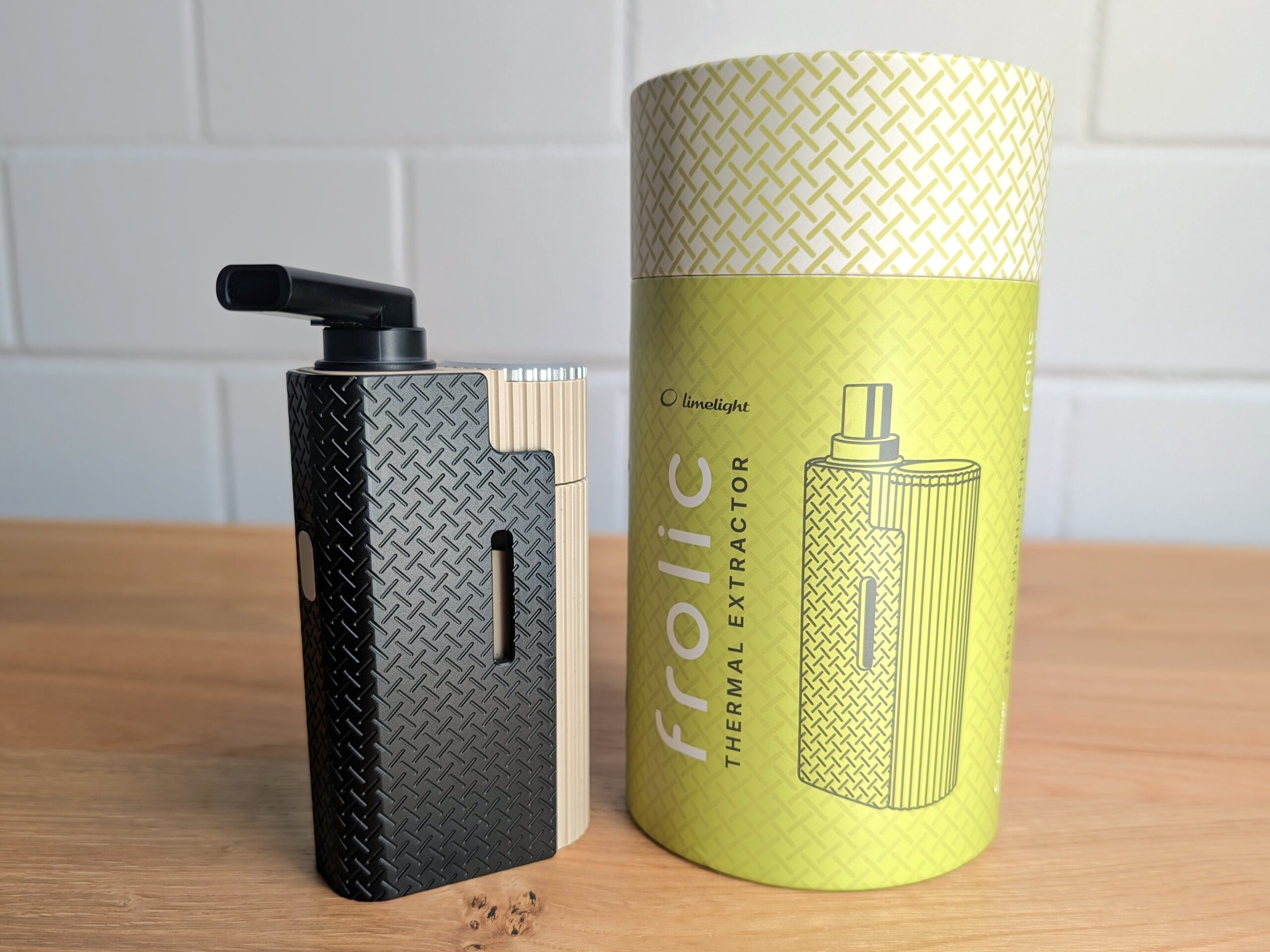 Der Frolic Thermal Extractor steht auf einem Holztisch. rechts daneben steht die zylinderförmige Pappverpackung Im Hintergrund ist eine unscharfe weiße Backsteinwand zu erkennen.