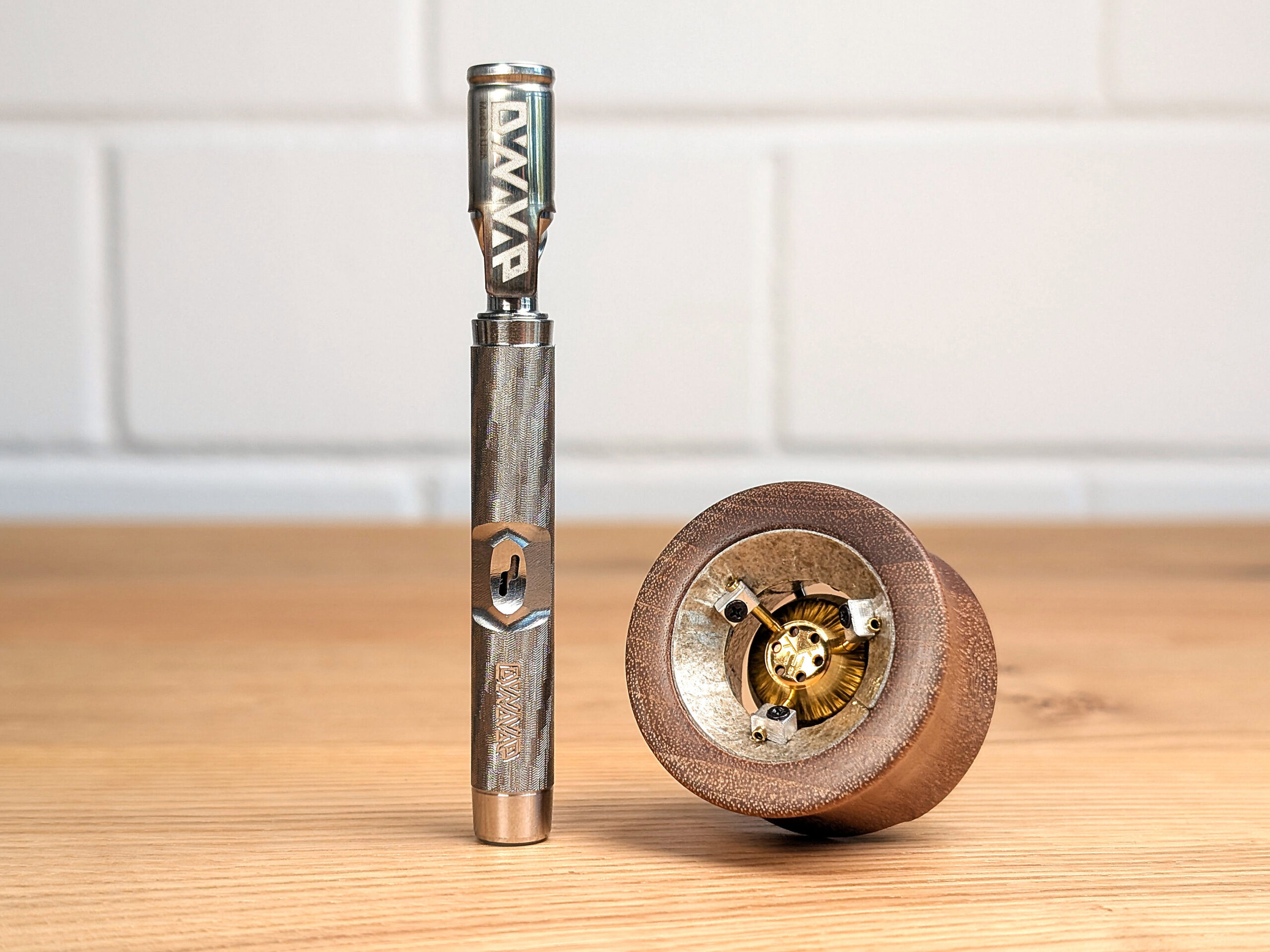 The DynaVap M7 and Vapman Vaporizer standing next to each other on a wooden table with a blurred white brick wall in the background.