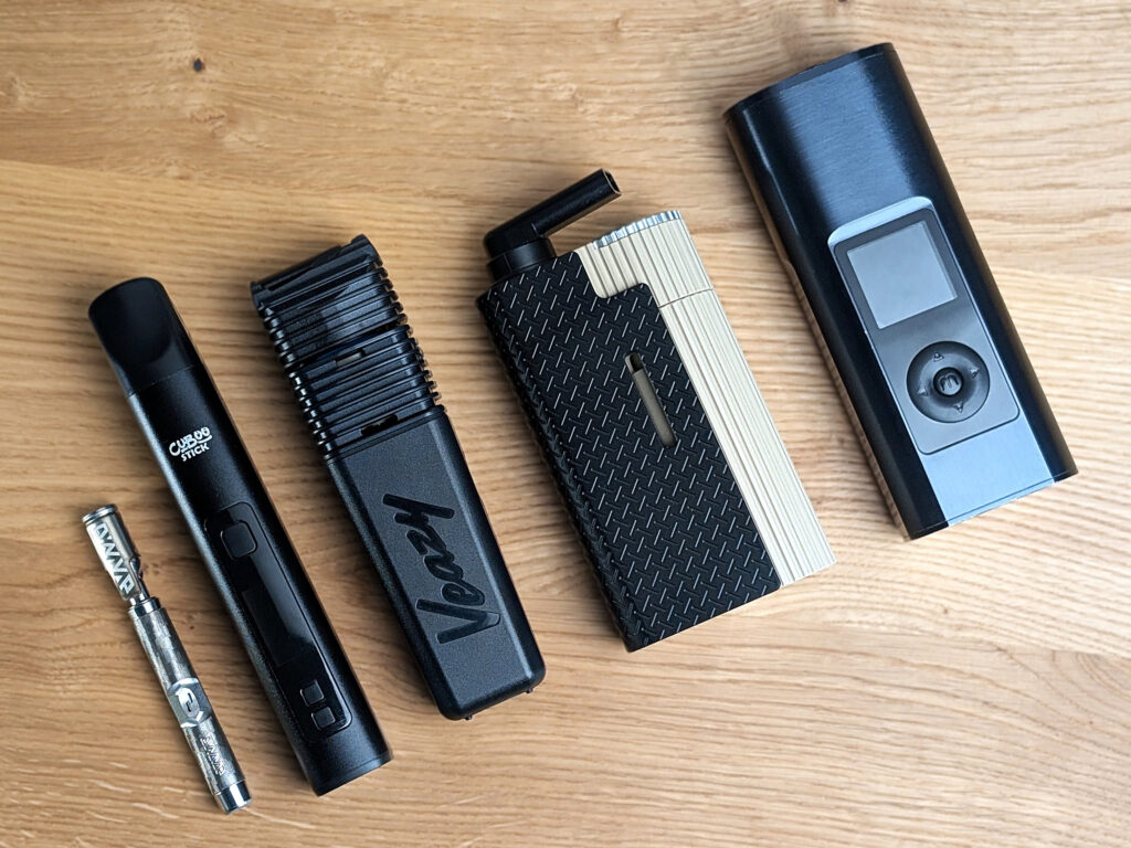 Fünf elektrische Vaporizer liegen auf einem Holztisch. Von links nach rechts: DynaVap G3, Cuboo Stick, Veazy, Frolic, Arizer Solo 3.