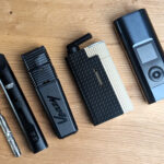 Fünf elektrische Vaporizer liegen auf einem Holztisch. Von links nach rechts: DynaVap G3, Cuboo Stick, Veazy, Frolic, Arizer Solo 3.