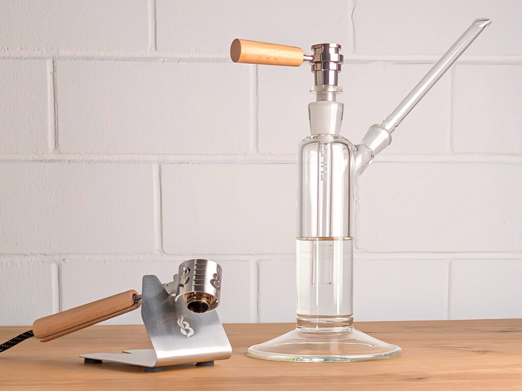 Ein Brennix Lift-Off Titan-Kopf steckt auf einem Ehle Power Tower Wasserfilter. Links daneben steht die Edelstahl-Halterung des Vaporizers mit eingraviertem Brennix Logo. In der Halterung ist der Brennix Lift-Off Vaporizer eingesetzt.