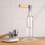 Ein Brennix Lift-Off Titan-Kopf steckt auf einem Ehle Power Tower Wasserfilter. Links daneben steht die Edelstahl-Halterung des Vaporizers mit eingraviertem Brennix Logo. In der Halterung ist der Brennix Lift-Off Vaporizer eingesetzt.