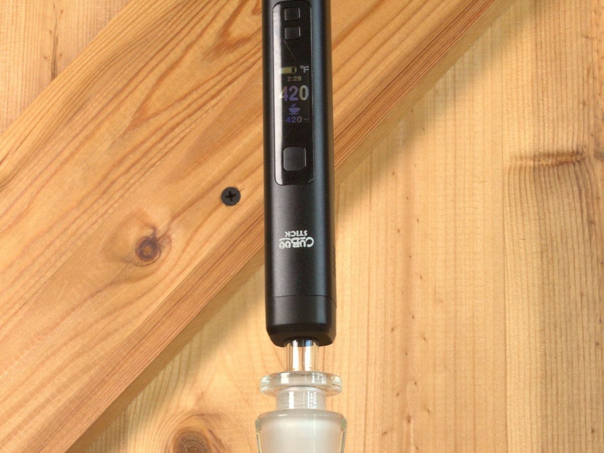 Ein schwarzer Cuboo Stick Vaporizer ist aufrecht an einen Glasadapter gesteckt und steht vor einer Holzwand mit diagonalen Holzleisten. Das Display zeigt 420 °F an und ist deutlich sichtbar.