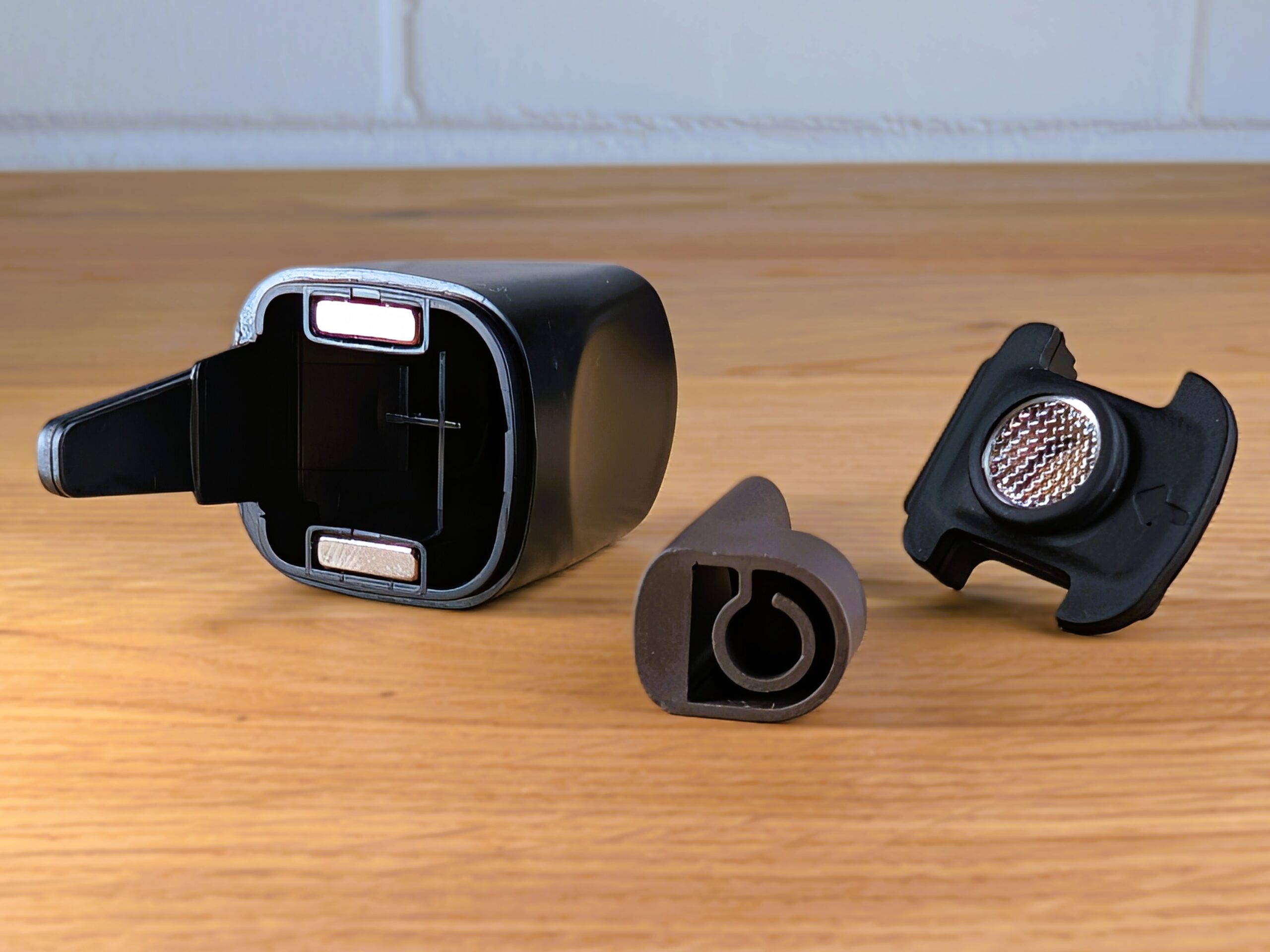 Ein auseinandergenommenes Mundstück des Cuboo Stick Vaporizers liegt auf einem Holztisch. Die drei Einzelteile – das leere Kunststoffgehäuse, das herausgenommene Keramik-Kühlelement und der Silikoneinsatz – sind deutlich voneinander getrennt platziert. Im Hintergrund ist eine unscharfe weiße Wand zu sehen.