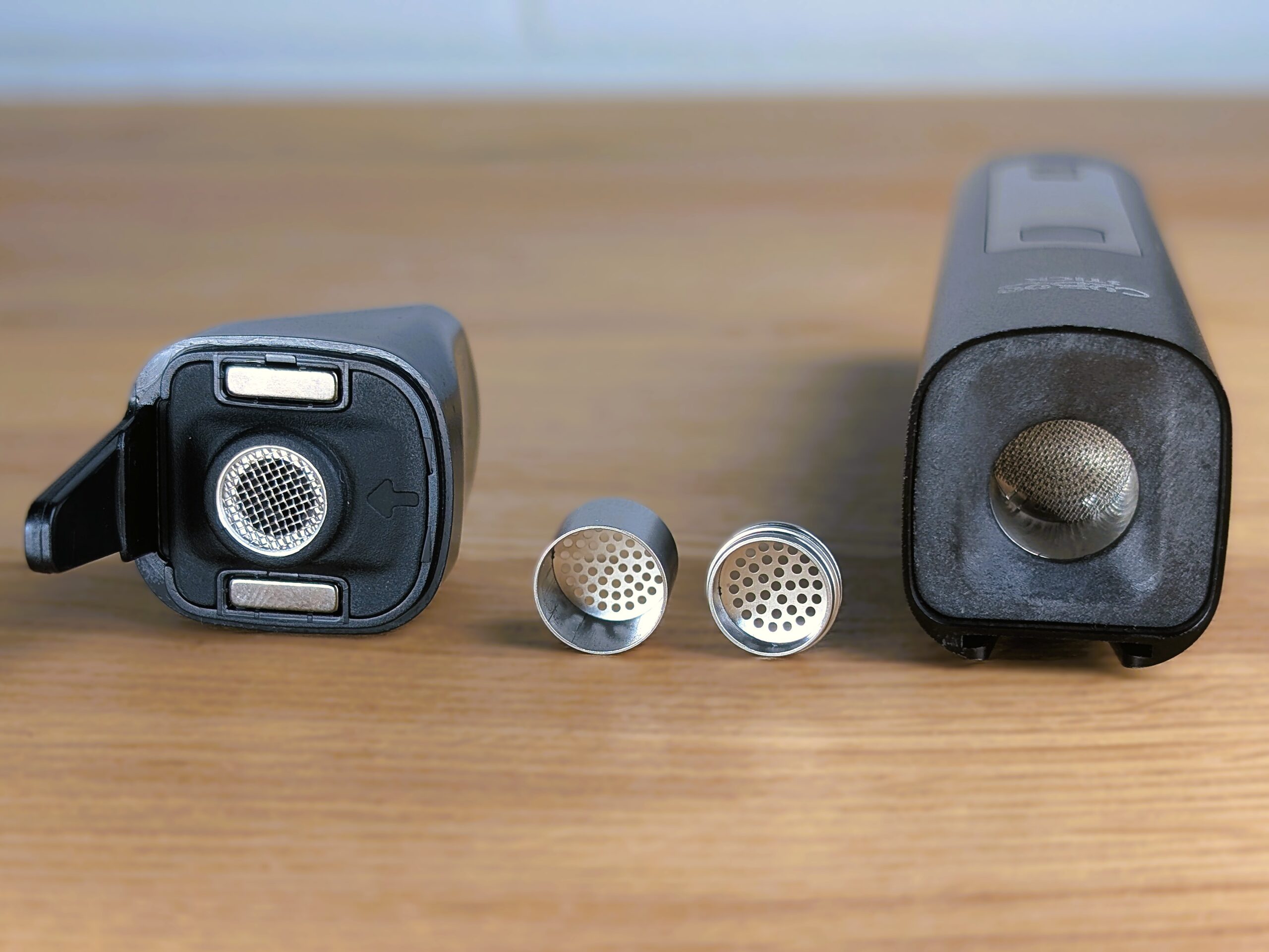 Der geöffnete Cuboo Stick Vaporizer liegt auf einem Holztisch, das Mundstück und die Heizkammer sind sichtbar. Eine geöffnete Edelstahlkapsel und der Deckel liegen davor, beide mit feinen Löchern. Im Hintergrund ist eine weiße Wand zu erkennen.