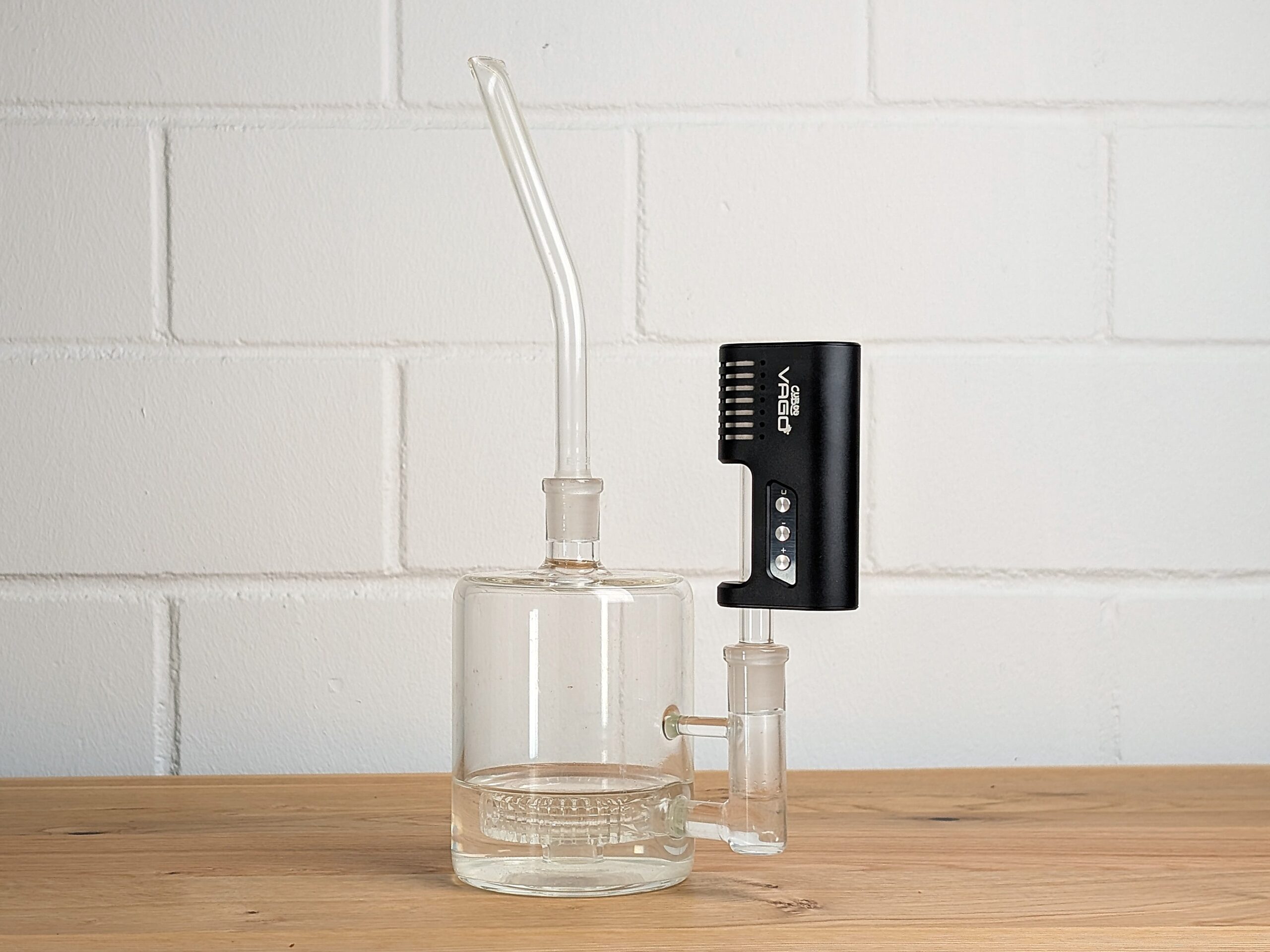 Ein Bubbler steht auf einem Holztisch. In den Bubbler ist der Cuboo VaGo Vaporizer eingesteckt. Im Hintergrund ist eine unscharfe weiße Backsteinwand zu erkennen.