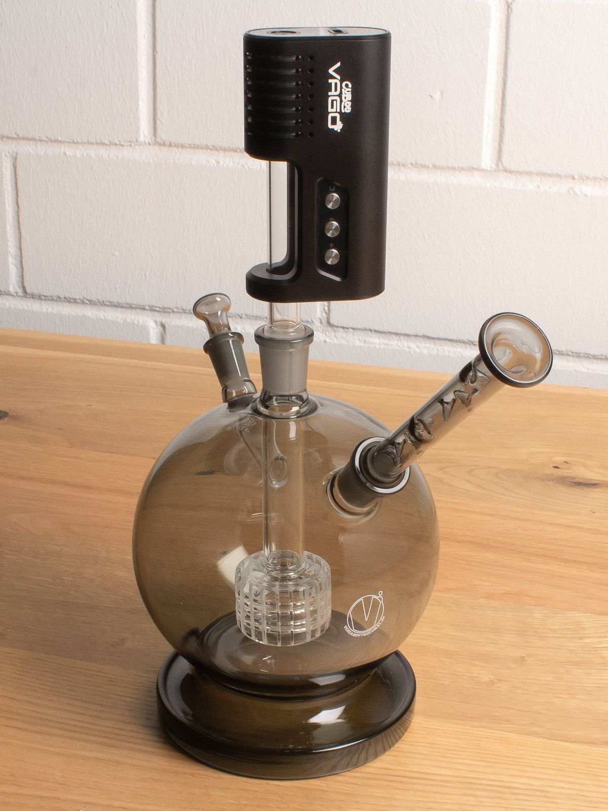 Ein Wasserfilter in Kugelform mit Trommelperkolator steht vor einer weißen Steinwand auf einem Holztisch. Im Wasserfilter ist der Cuboo-Vago-Vaporizer von oben eingesteckt.