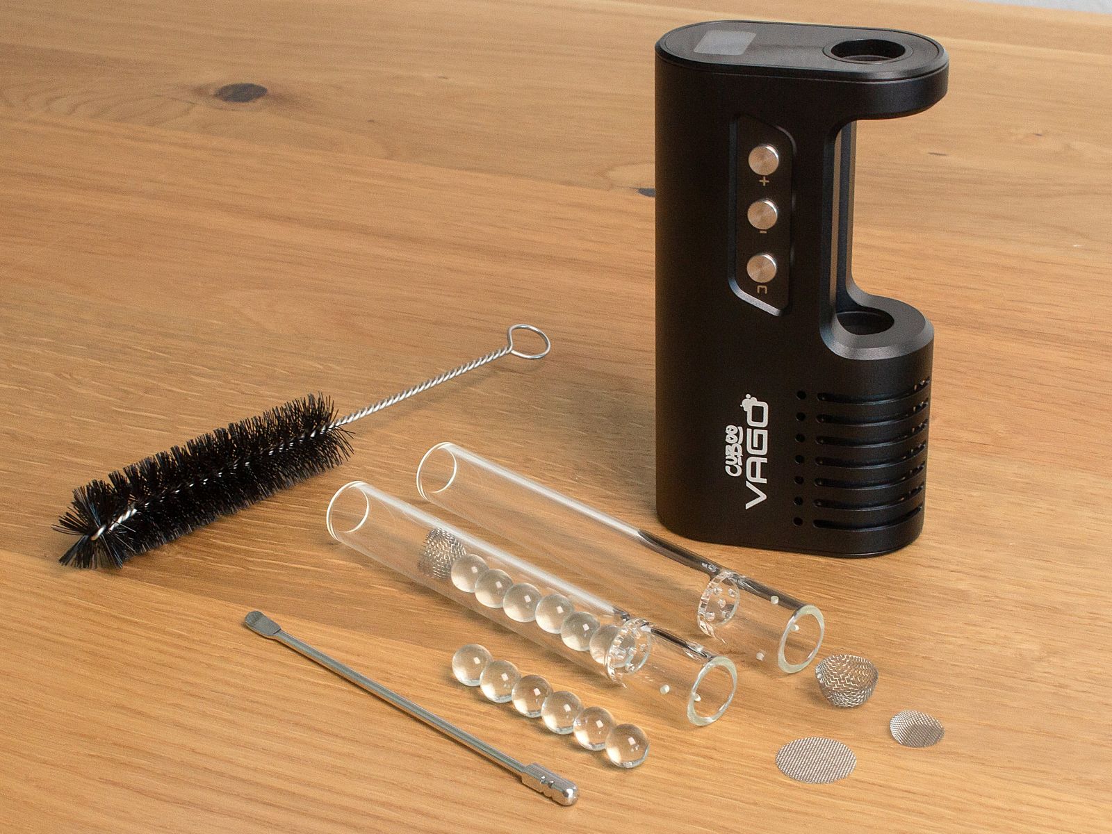 Der Cuboo-Vago-Vaporizer steht auf einem Tisch. Daneben liegen eine Reinigungsbürste, ein Edelstahl-Werkzeug, drei Siebe, sechs Glaskugeln und zwei Mundstücke. Eins der Mundstücke ist mit sechs Glaskugeln gefüllt und mit einem Sieb verschlossen.