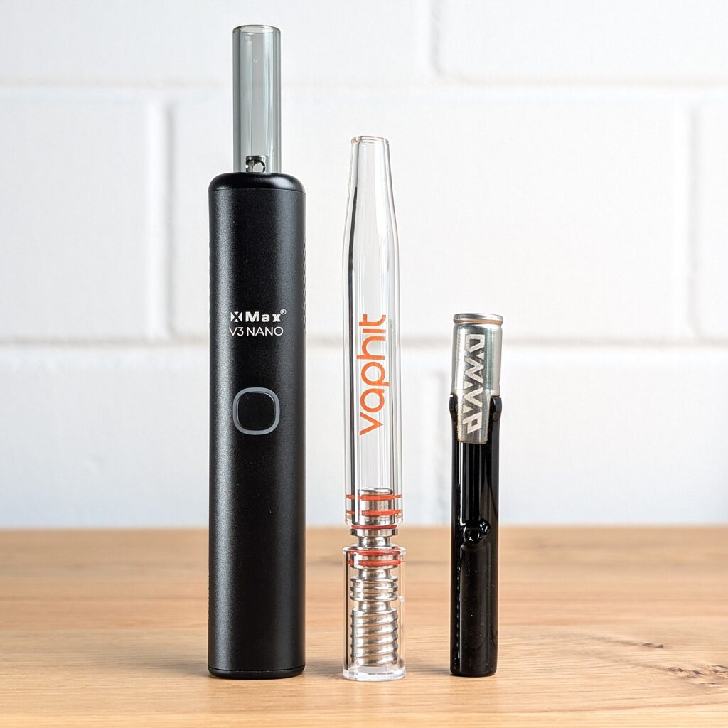 Ein XMAX Nano V3, ein VapHit ECO und ein Dynavap G3 Vaporizer stehen nebeneinander auf einem Holztisch. Im Hintergrund ist eine unscharfe weiße Backsteinwand zu erkennen.
