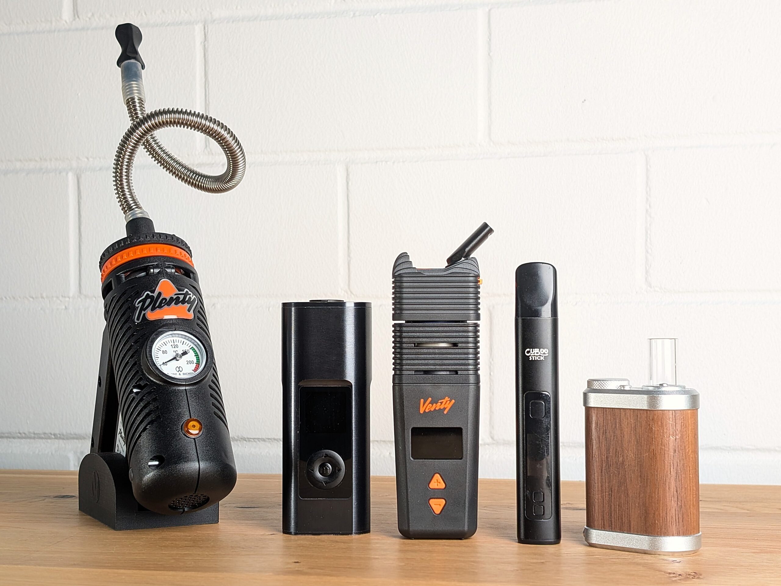 Five different vaporizers are lined up on a wooden table. From left to right: Plenty, Arizer Solo 3, Venty, Cuboo Stick, Tiny Might 2