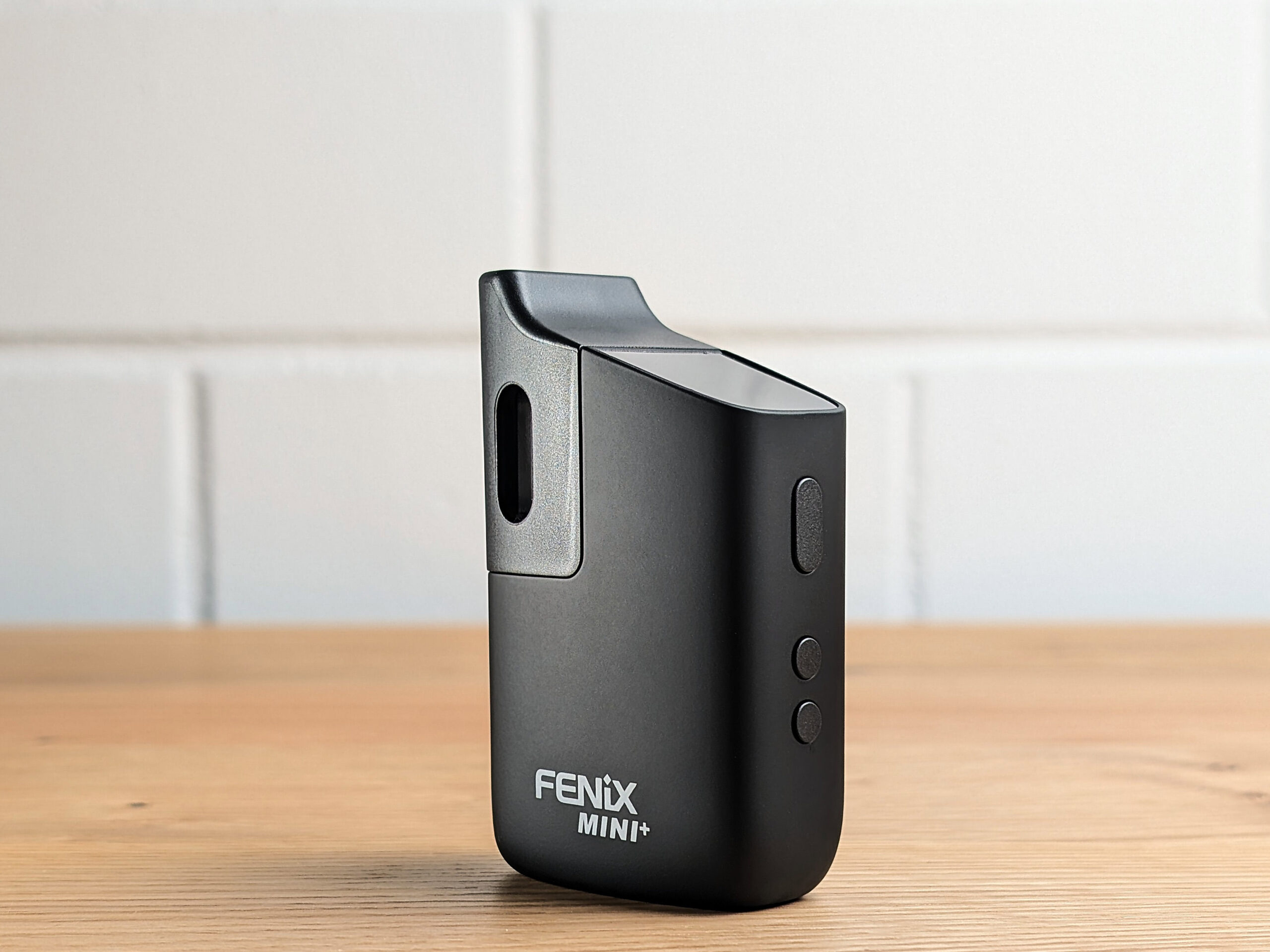 The Fenix Mini Plus vaporizer stands at an angle on a wooden table. The buttons on the narrow side are facing the viewer. A blurred white brick wall is visible in the background.