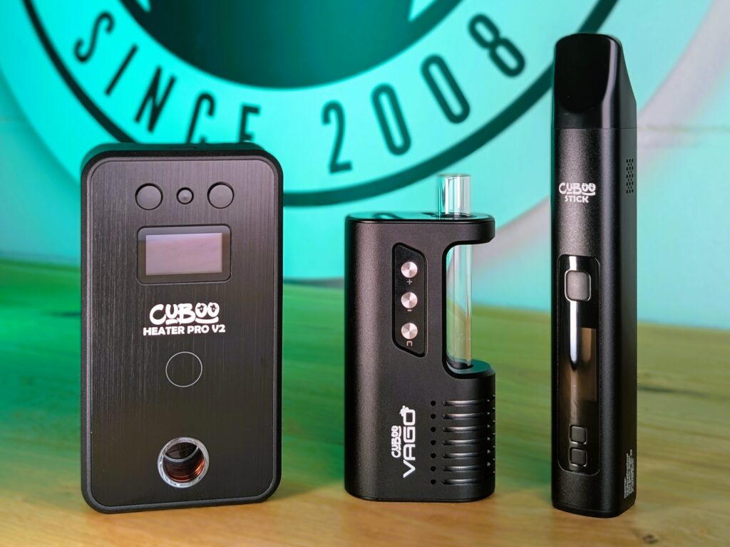Ein Cuboo Heater Pro V2, ein Cuboo-VaGo-Vaporizer und ein Cuboo-Stick-Vaporizer stehen nebeneinander auf einem Holztisch. Im Hintegrund ist ein Teil des Verdampftnochmal-Logos ist im Hintergrund zu erkennen.