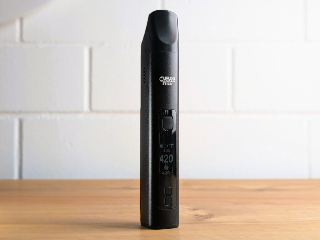 Ein schwarzer tragbarer Vaporizer mit der Aufschrift ‚Cuboo Stick‘ steht aufrecht auf einem hellen Holztisch. Das Gerät zeigt eine digitale Temperaturanzeige von 420 Grad Fahrenheit. Im Hintergrund ist eine weiße Backsteinwand.
