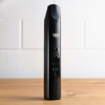 Ein schwarzer tragbarer Vaporizer mit der Aufschrift ‚Cuboo Stick‘ steht aufrecht auf einem hellen Holztisch. Das Gerät zeigt eine digitale Temperaturanzeige von 420 Grad Fahrenheit. Im Hintergrund ist eine weiße Backsteinwand.