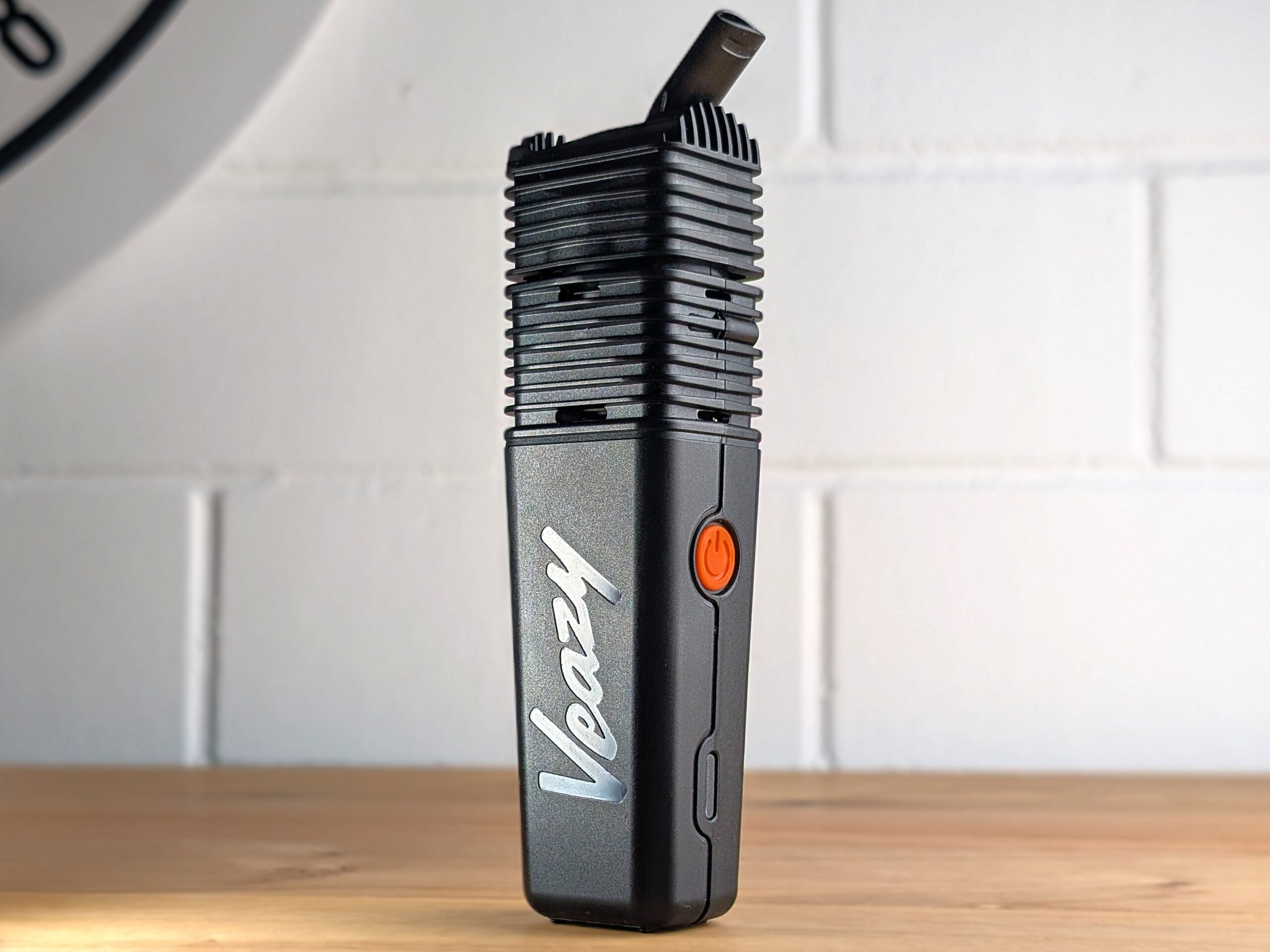 The Veazy vaporizer sits on a wooden table. In the background, a blurry white brick wall can be seen.