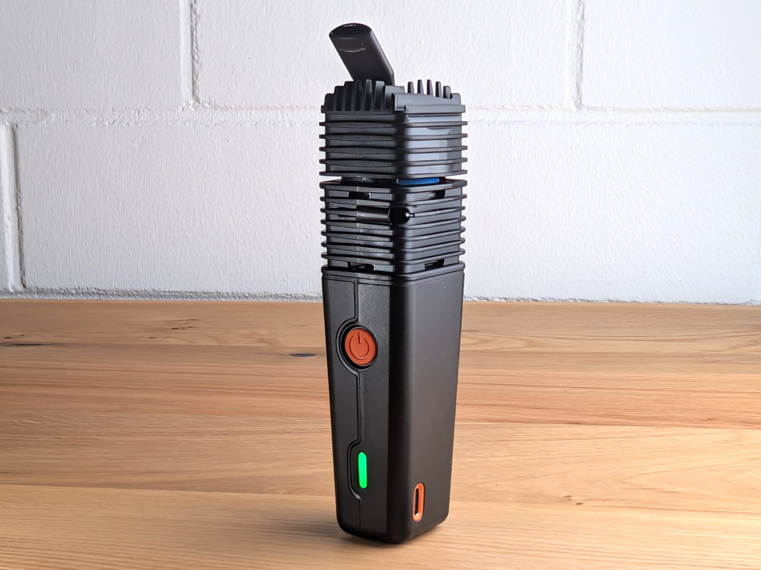 The Veazy vaporizer sits on a wooden table. In the background, a blurry white brick wall can be seen.