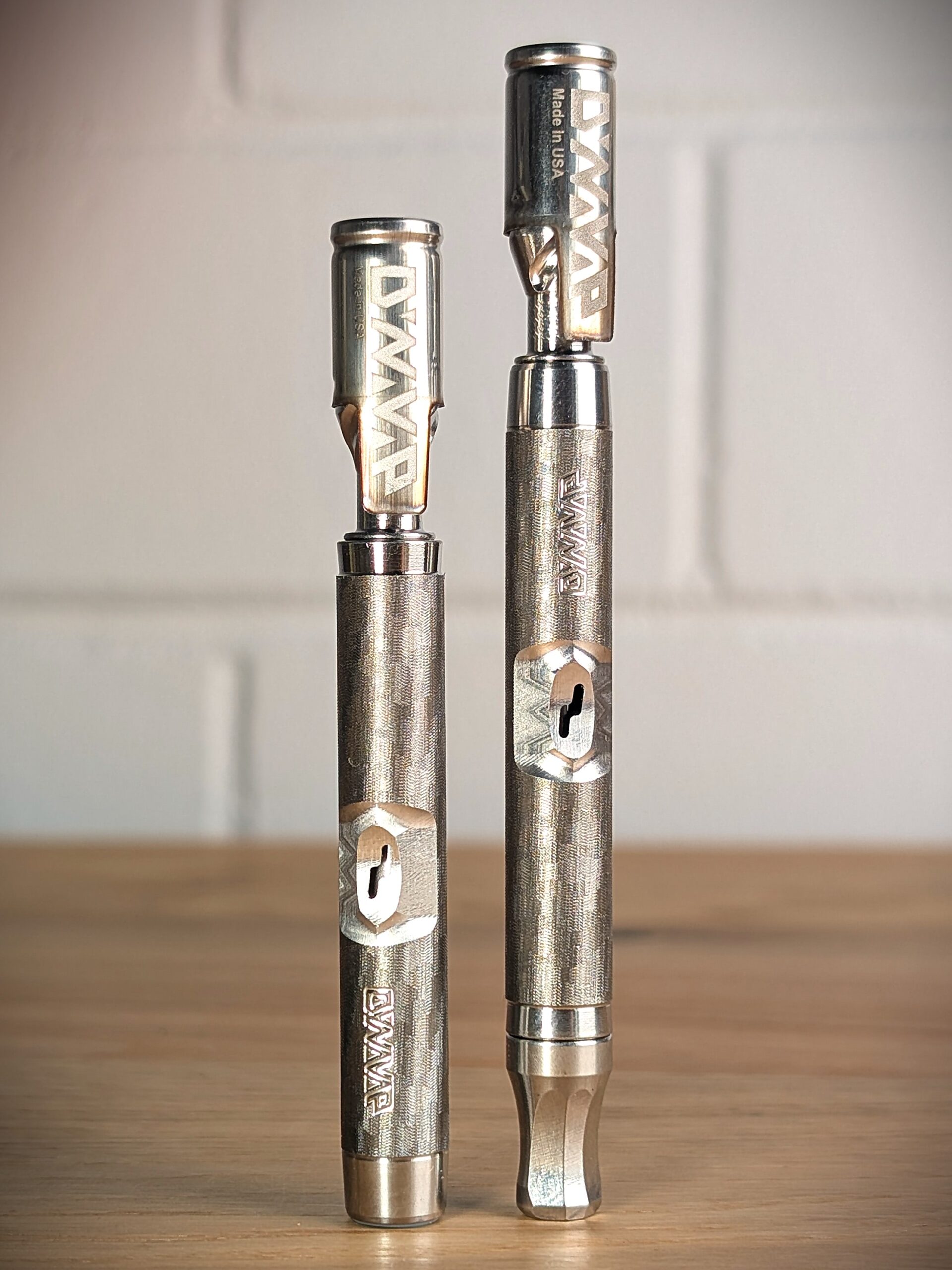 Zwei Dynavap Vaporizer stehen nebeneinander auf einem Holztisch. Das linke Model ist ein Dynavap M7, das rechte ein Dynavao M7XL. Im Hinztergrund ist eine weiße Backsteinwamd unscharf zu erkennen.