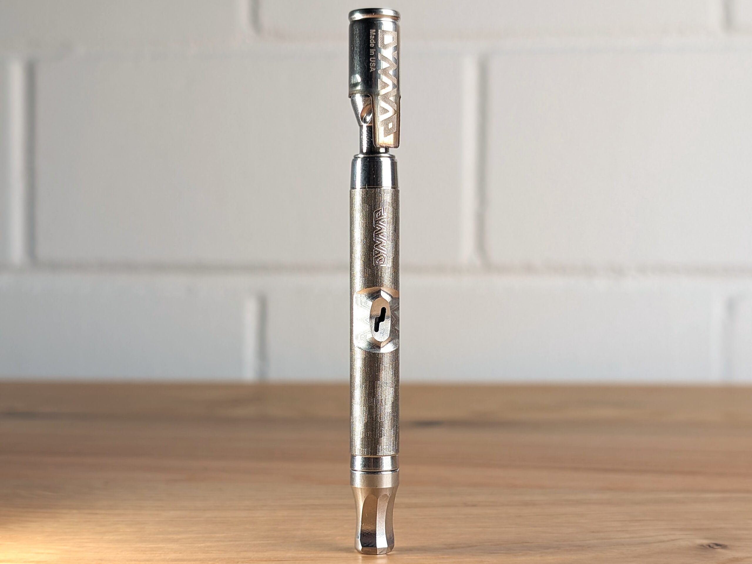The Dynavap vaporizer stands on a wooden table. A blurred white brick wall is visible in the background.