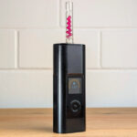 Der Arizer Solo 3 Vaporizer steht auf einem Holztisch. Ein gläserenes Mundstück, das mit kleinen Ruby-Balls gefüllt ist, steckt von oben im Vaporizer. Im Hintergrund ist eine unscharfe weiße Backsteinwand zu erkennen.