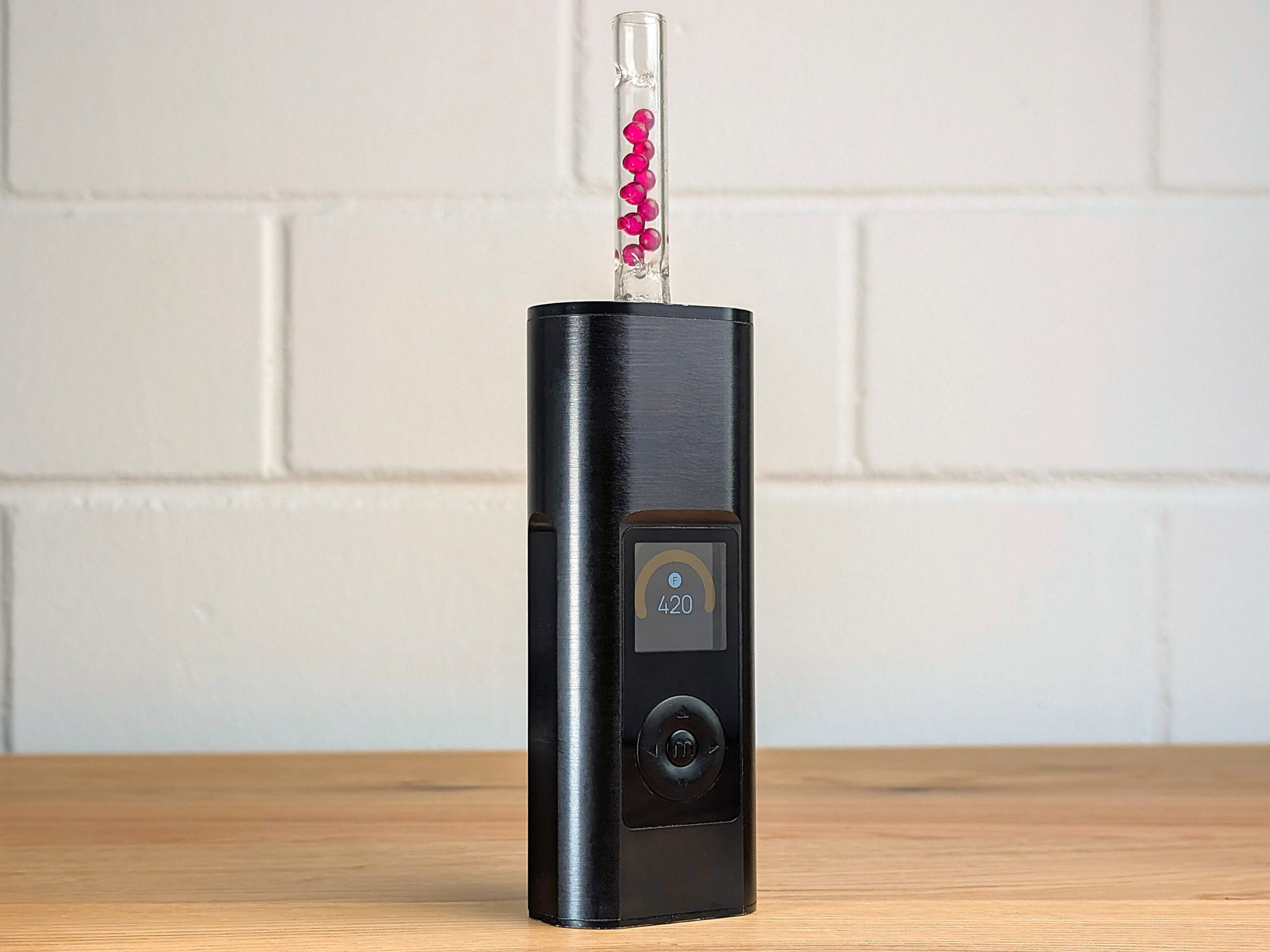 Der Arizer Solo 3 Vaporizer steht auf einem Holztisch. Ein gläserenes Mundstück, das mit kleinen Ruby-Balls gefüllt ist, steckt von oben im Vaporizer. Im Hintergrund ist eine unscharfe weiße Backsteinwand zu erkennen.