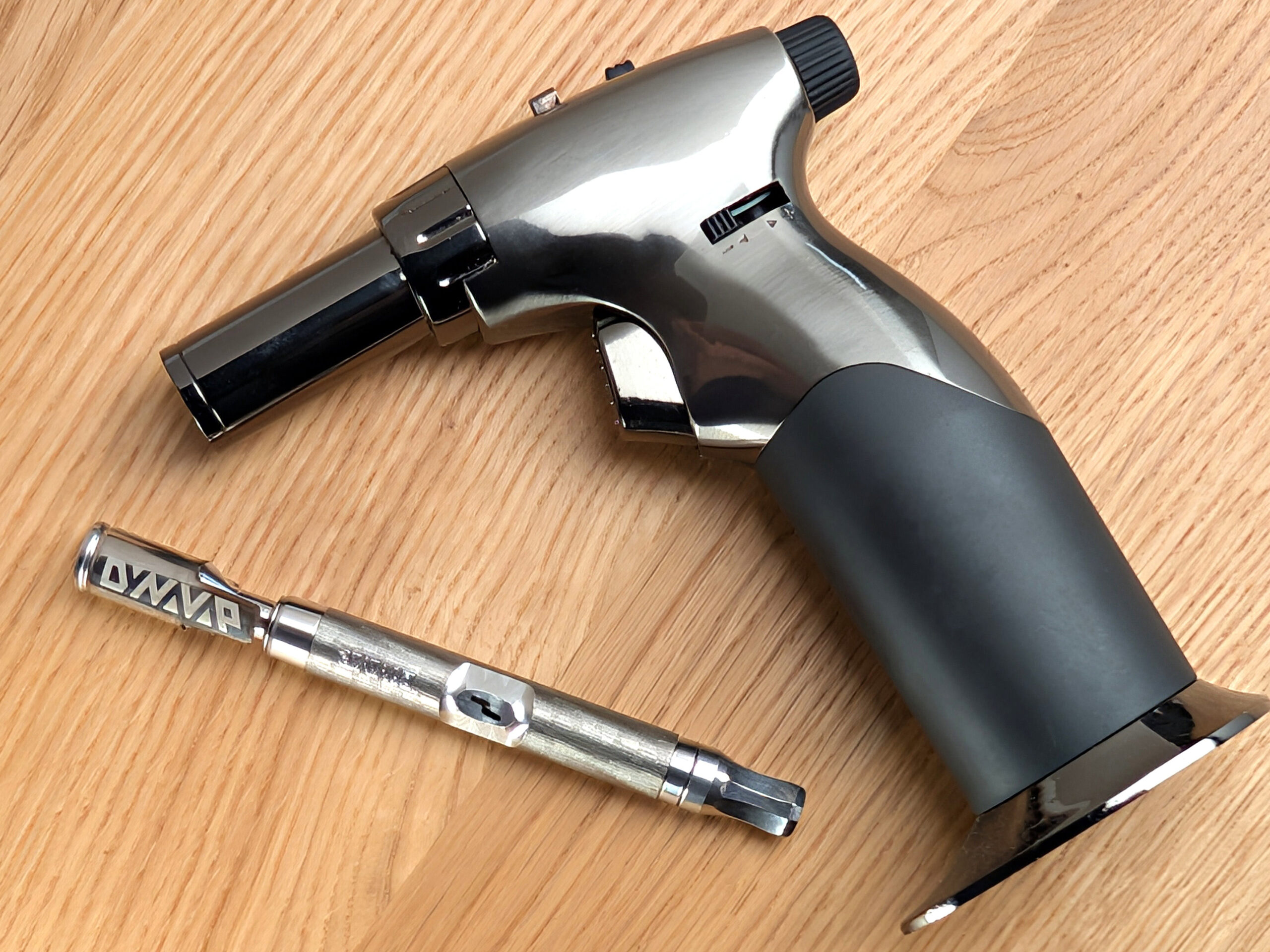 A DynaVap M7 vaporizer and a jet-flame lighter lie angled next to each other on a wooden table.