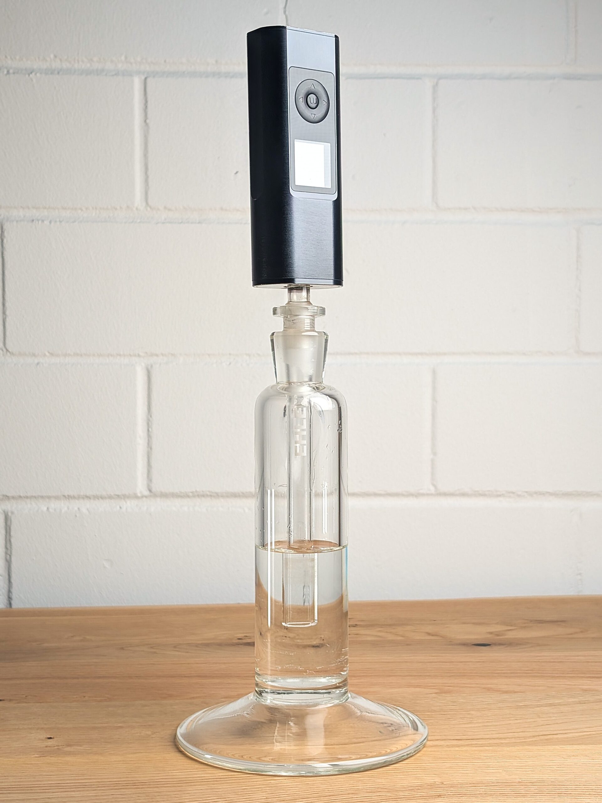 Arizer Solo 3 vaporizer sits on a Power Tower glass water filter filled with water on a wooden table in front of a white brick wall.