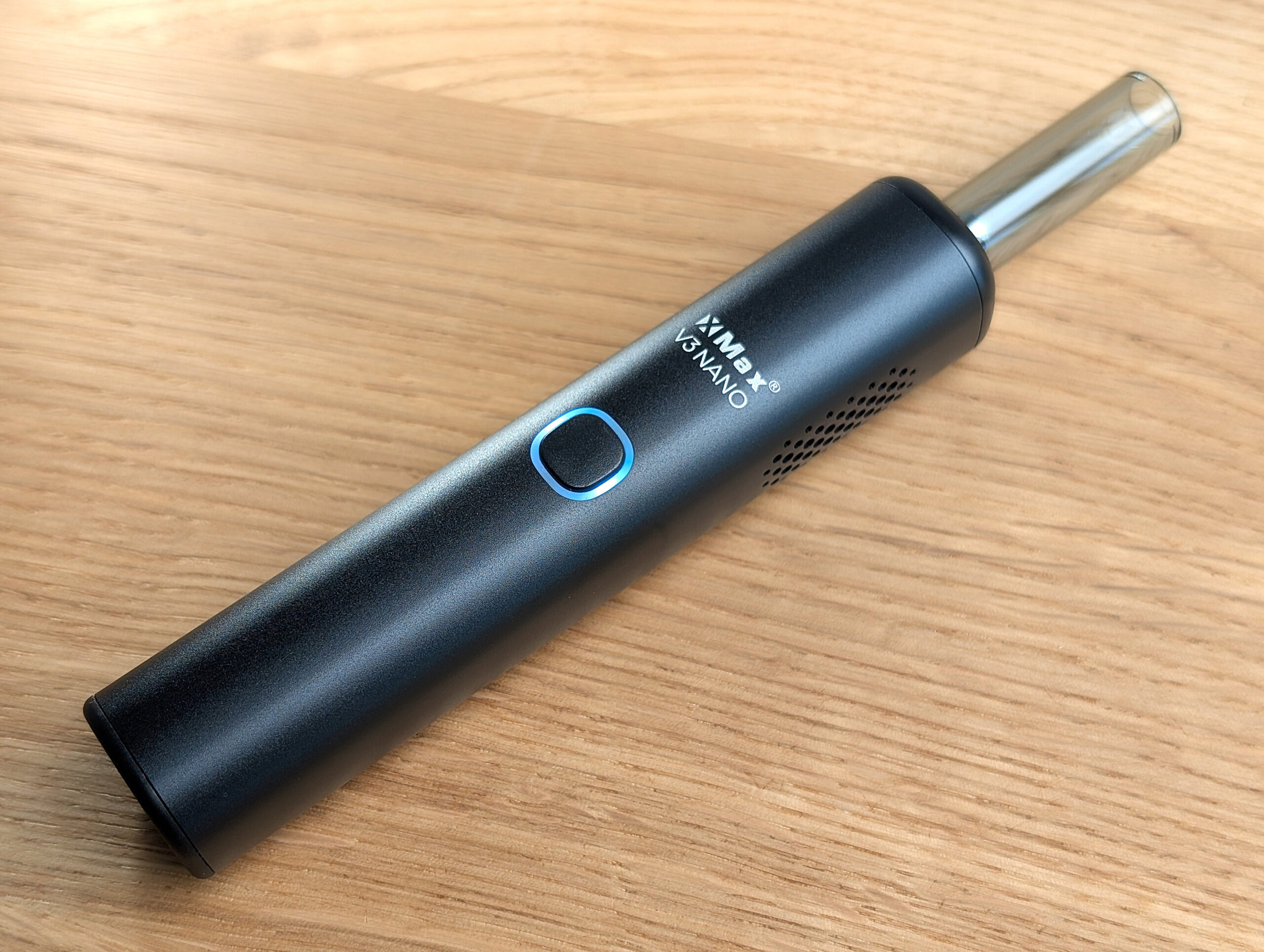 An XMAX V3 Nano vaporizer lies at a 45° angle on a wooden table. The status LED is glowing blue.