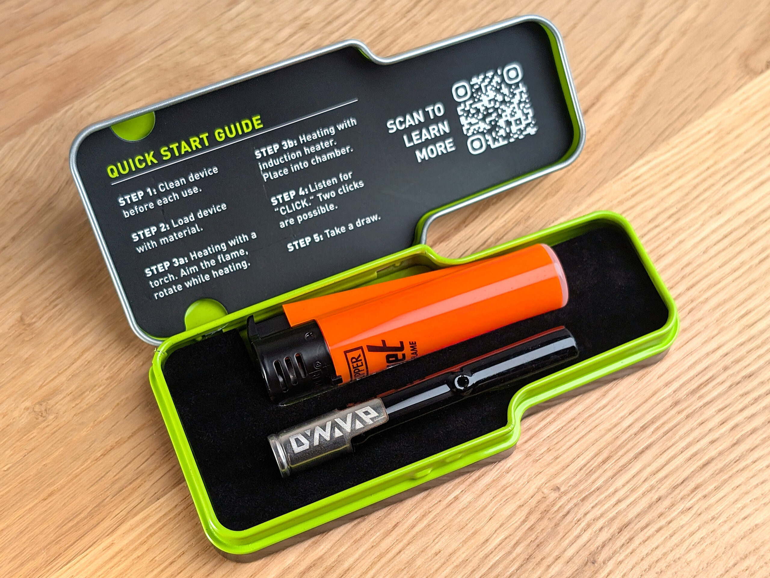 An open DynaVap tin case on a wooden table. Inside are an orange Clipper jet flame lighter and a black DynaVap G3 vaporizer.