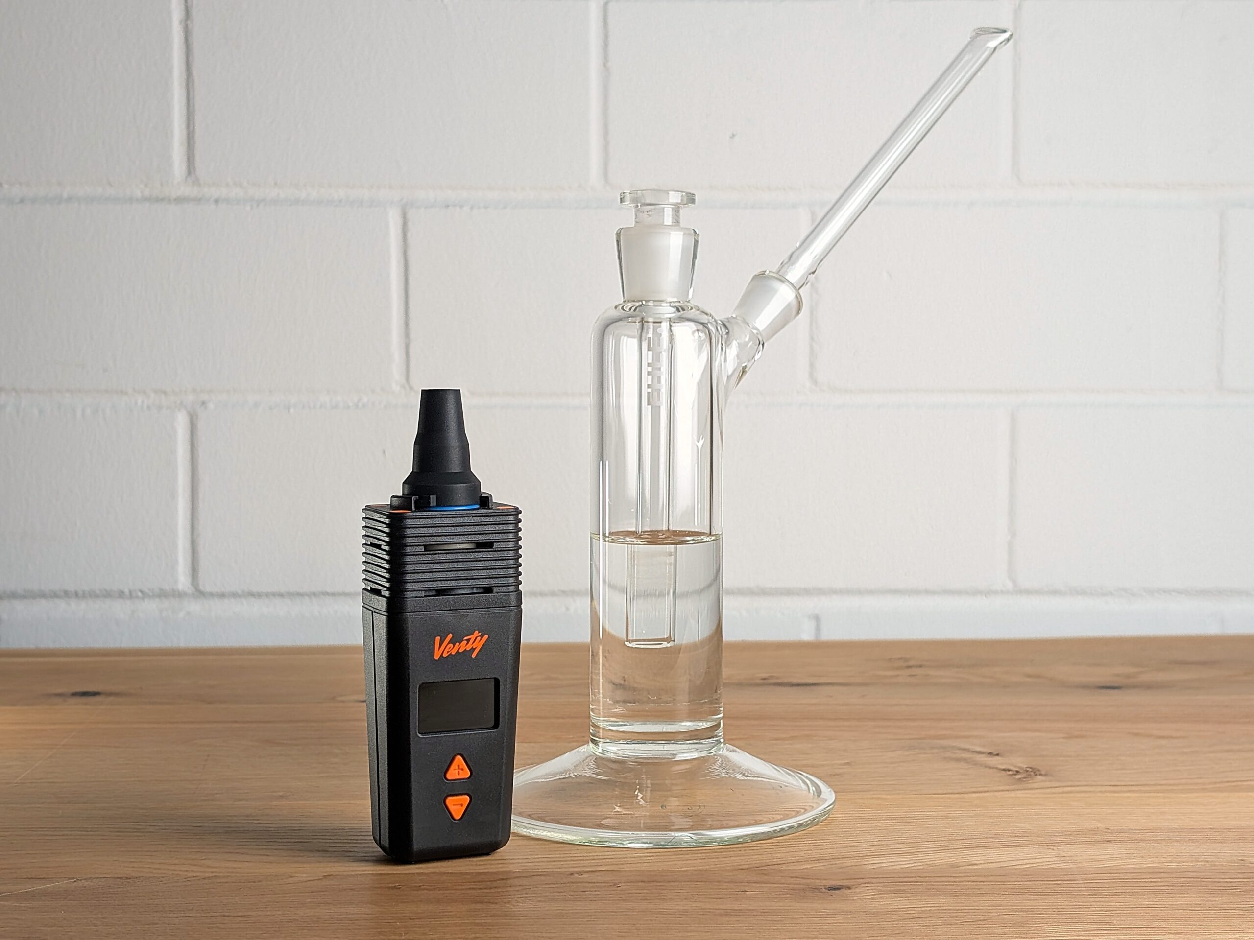 A Venty vaporizer sits on a wooden table. Instead of the cooling element, a WPA is attached to the Venty. To the right, a Power Tower water filter with a straight Ehle mouthpiece is visible. In the background is a blurred brick wall.