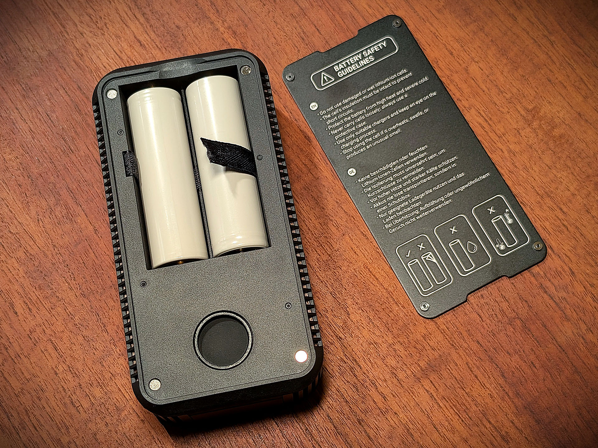 The Cuboo Heater XL is lying on a wooden table with the battery cover open. The battery cover is lying to the right of it. Safety instructions can be seen on the inside of the battery cover.