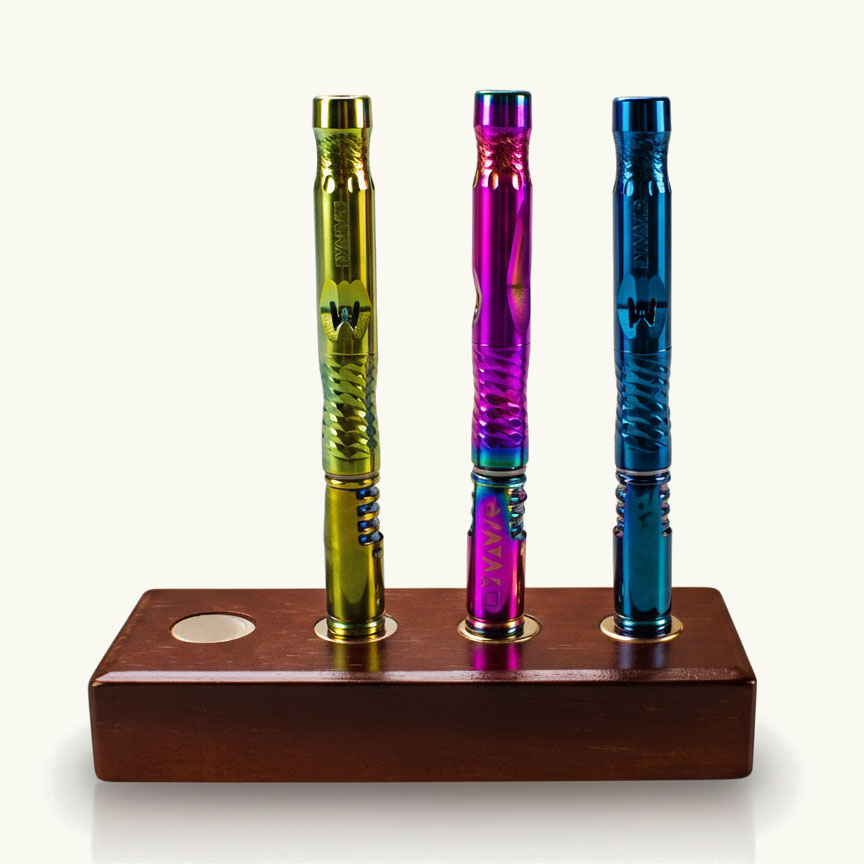 Drei farbenfroh schimmernde Vaporizer (Gold/Gelb, Violett/Blau, Blau) stehend in einem rechteckigen Halter aus dunklem Holz.