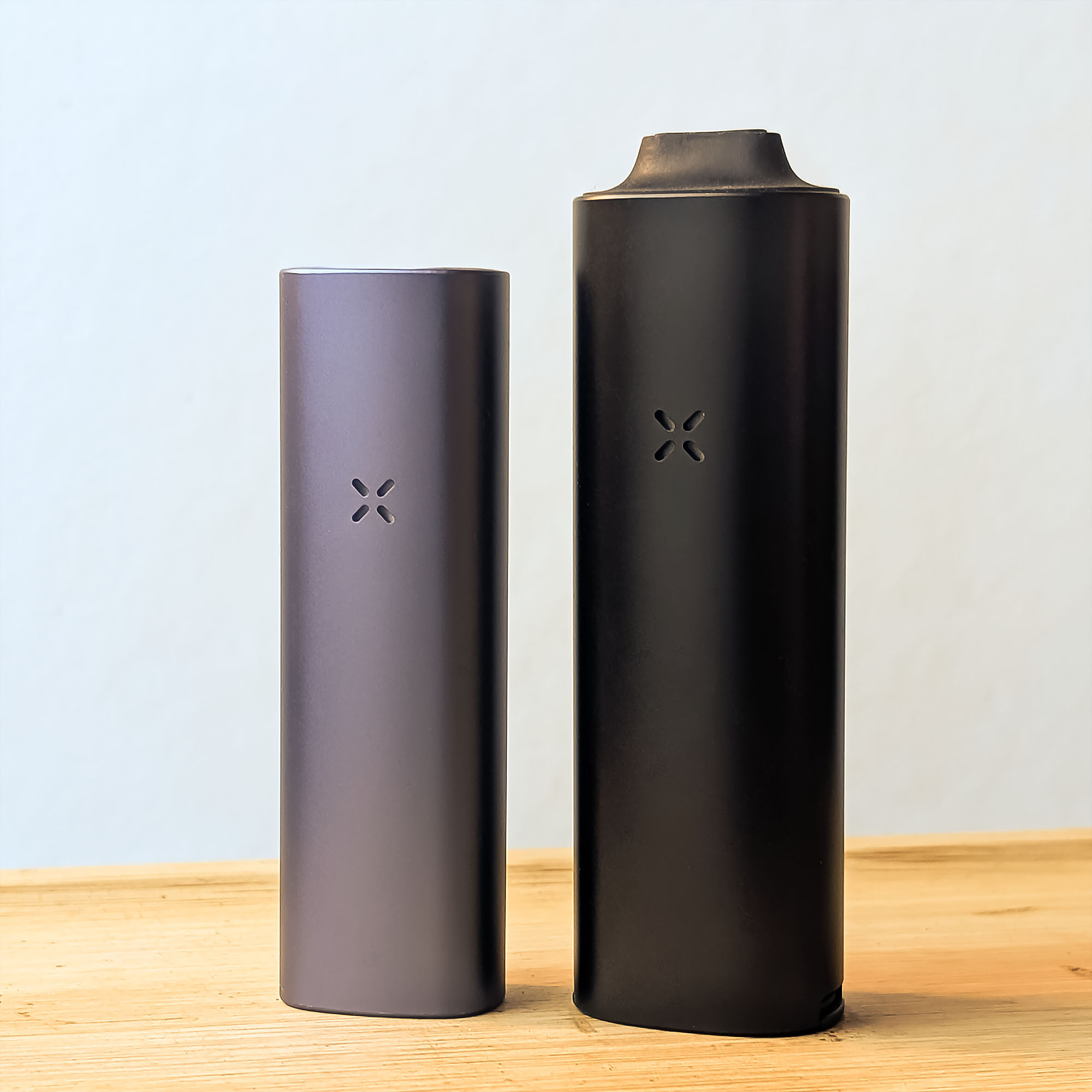 Ein direkter Größenvergleich zwischen dem PAX Plus Vaporizer und dem neuen, etwas größeren PAX FOUR, beide stehend auf einem hellen Holztisch.