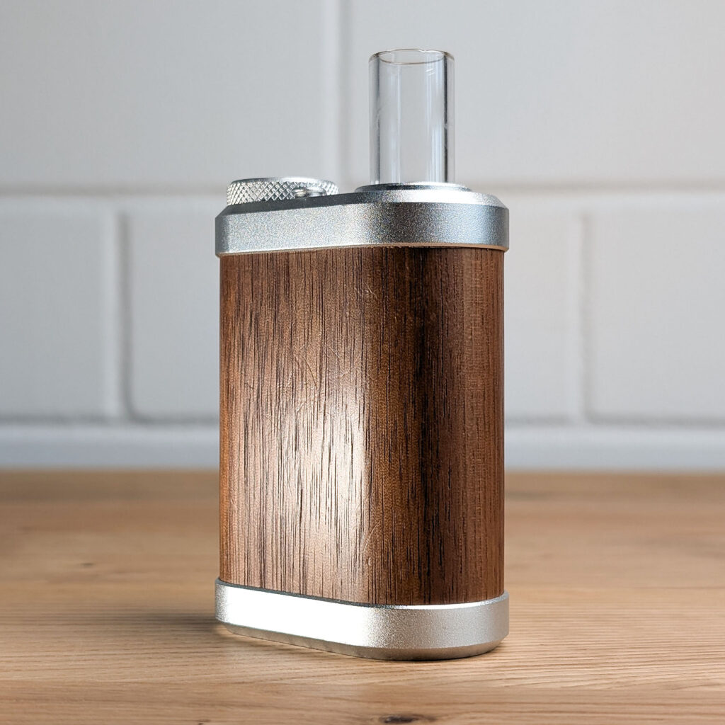 The Tiny Might 2 vaporiser is sitting on a wooden table. In the background, a blurred white brick wall can be seen.