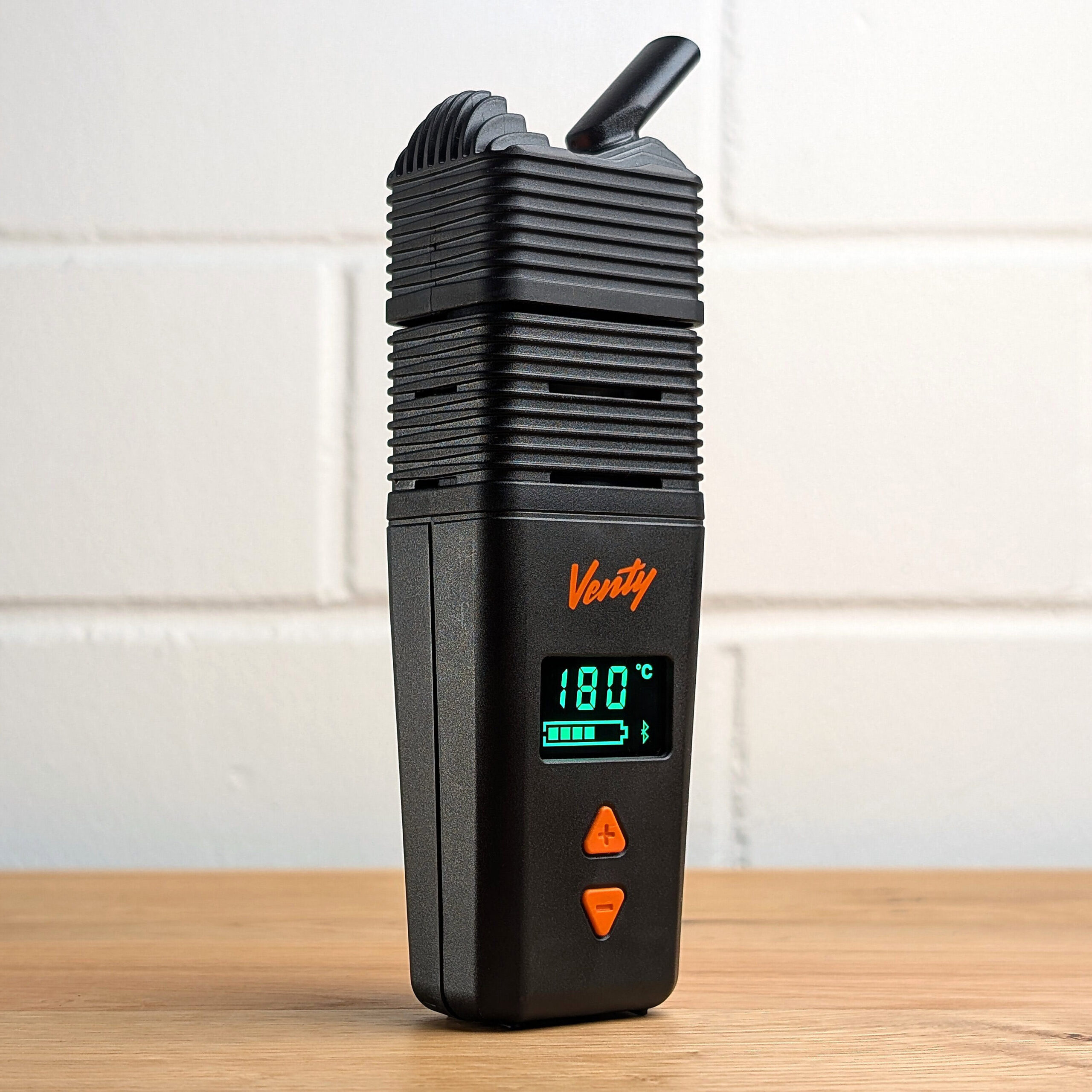 Auf einem Holztisch steht schräg ein Venty Vaporizer. Das Display leuchtet grün und zeigt die Temperatur, den Ladezustand und ein Bluetooth Symbol an. Im Hintergrund ist eine unscharfe weiße Backsteinwand zu erkennen.